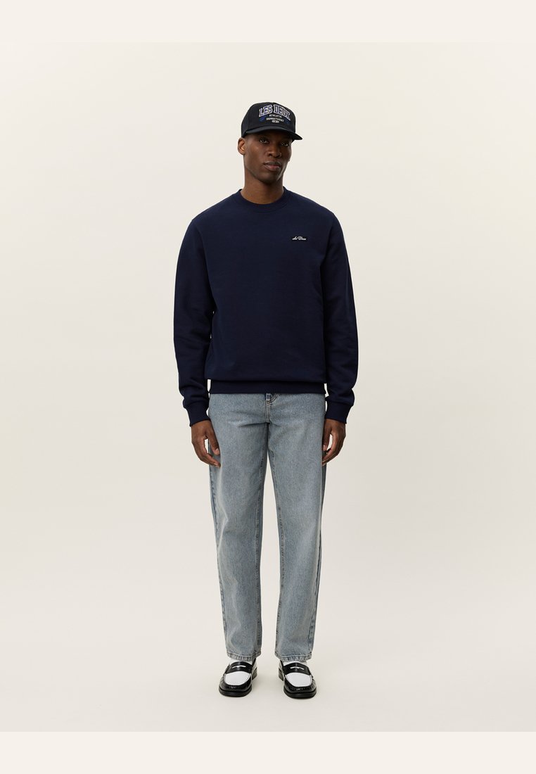 Marineblå genser med rund hals, kombinert med lyseblå denimjeans. Svart og hvite sko, og en mørk caps med tekstlogo.