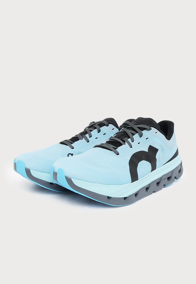 Un par de zapatillas de running azul claro con cordones negros, grandes logotipos negros en los laterales y suelas grises gruesas, sobre un fondo liso.