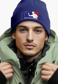 Blue knitted beanie featuring a white and red logo patch that reads "MAJOR LEAGUE BEERPONG," worn with a green puffer jacket.