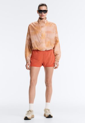 Femeie purtând un windbreaker tie-dye supradimensionat, culoare piersică, pantaloni scurți de culoare ruginie, șosete albe, adidași bej și ochelari de soare negri supradimensionați, stând pe un fundal alb.