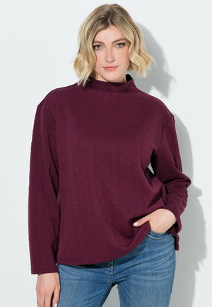 Kvinde med kort blondt hår iført en struktureret bordeaux-langærmet top og blå jeans med den ene hånd i lommen mod en hvid baggrund.