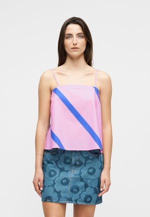 Νεαρή γυναίκα που φοράει ροζ τοπάκι με μπλε διαγώνιες ρίγες και μπλε τζιν φούστα με μεγάλα λουλουδάτα σχέδια, στέκεται σε λευκό φόντο.