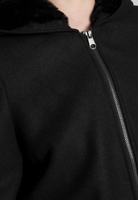 Sudadera negra con cremallera hecha de material de forro polar, que presenta una capucha forrada de piel sintética y detalles en cremallera plateada. Textura suave con un corte relajado.