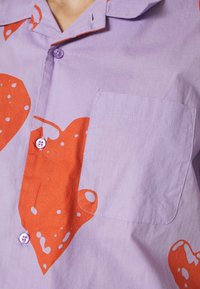 Tričko s krátkým rukávem v světle fialové barvě z bavlny, se velkými červenými srdcovými vzory, kapsou na levé straně a fialovými knoflíky.