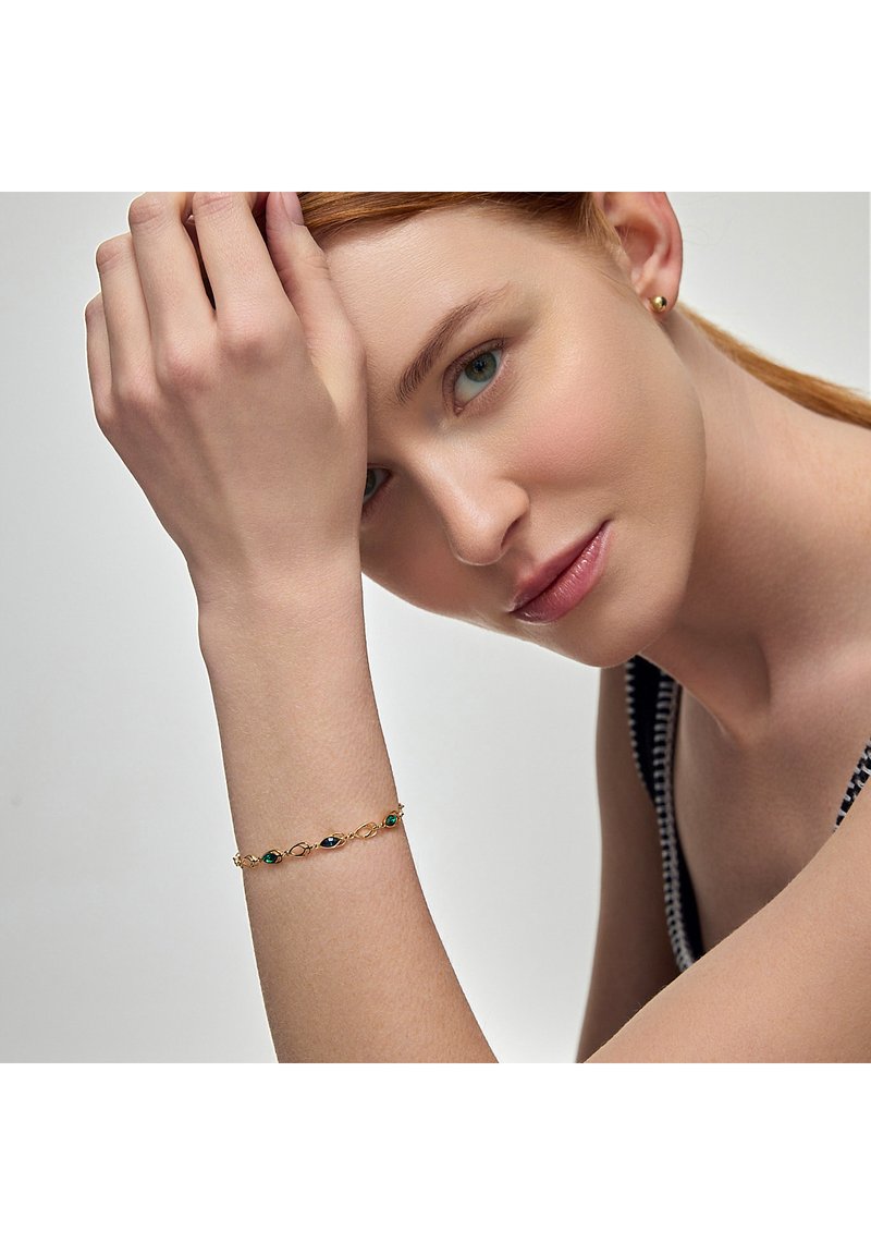 Złota bransoletka z zielonymi i niebieskimi kamieniami szlachetnymi w układzie naprzemiennym, noszona na nadgarstku, na neutralnym tle.