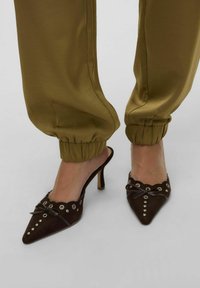 Mules à talons hauts en daim marron avec des orteils pointus, des bords festonnés et des détails métalliques en œillet, associés à un pantalon olive à revers.