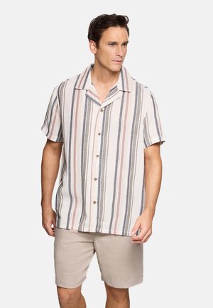 Hombre vistiendo una camisa de manga corta con botones y rayas verticales en beige, rosa y azul marino, combinada con pantalones cortos beige claro, de pie y mirando hacia la derecha.