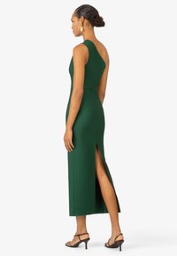 Robe midi sans manches verte avec une fente latérale montrant la jambe. Fabriquée dans un tissu lisse avec une silhouette ajustée. Associée à des talons noirs à lanières.