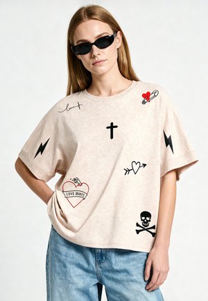 Vrouw draagt beige oversized T-shirt met tatoeages en symbolen waaronder hart, kruis, schedel en bliksemschichten, gecombineerd met blauwe jeans en zwarte zonnebril.