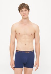 Navy blue Nike boxer briefs feature an orange branded waistband. The fabric appears smooth and fitted, with a mid-length cut.