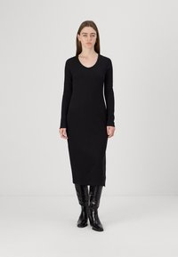 Vestido preto de manga longa com decote em V, silhueta ajustada, comprimento até à metade da panturrilha, feito de um tecido suave. Botas pretas até ao joelho com brilho.