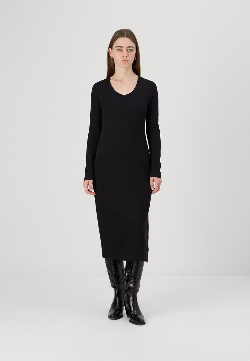 Vestido preto de manga longa com decote em V, silhueta ajustada, comprimento até à metade da panturrilha, feito de um tecido suave. Botas pretas até ao joelho com brilho.