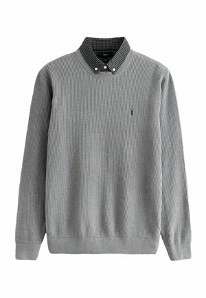 Pull en maille côtelée gris pour homme superposé sur un col de chemise à carreaux noir et blanc, manches longues, petit logo brodé sur la poitrine.