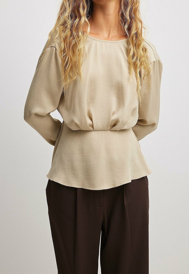 Femme aux longs cheveux blonds ondulés, portant un chemisier beige ample à manches longues et un pantalon marron foncé, debout les mains derrière le dos.