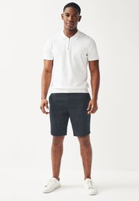 Polo bianco con colletto a zip, abbinato a pantaloni corti blu scuro e scarpe da ginnastica bianche. L'outfit presenta un design semplice e informale.