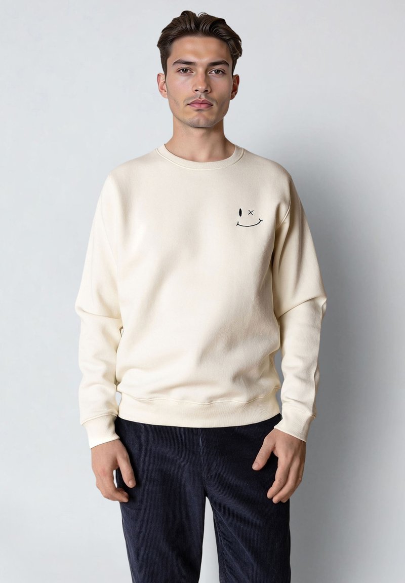 Homme portant un sweat-shirt crème avec un petit smiley noir faisant un clin d'œil sur la poitrine et un pantalon en velours côtelé foncé, debout devant un fond uni.