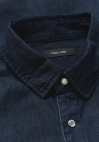 Camisa de mezclilla azul marino con cuello abotonado, que presenta una etiqueta negra con la inscripción "Matinique" y botones de color claro.