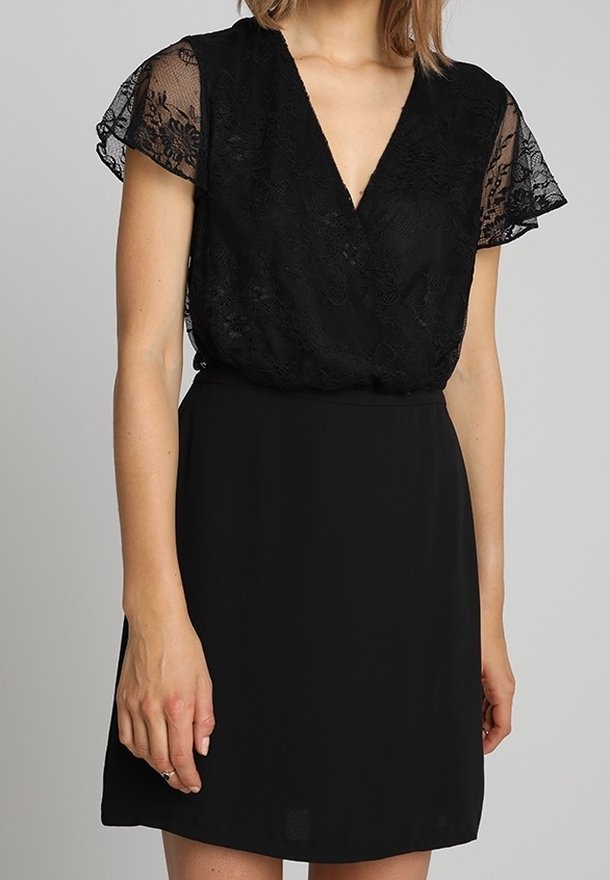 Femme portant une robe courte noire avec des manches courtes en dentelle et un profond décolleté en V, debout devant un fond gris uni.