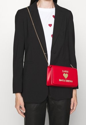 Kvinde iført sort blazer og bukser, hvid top med røde hjerte detaljer, bærende på en rød Love Moschino håndtaske med guld kædestrop.