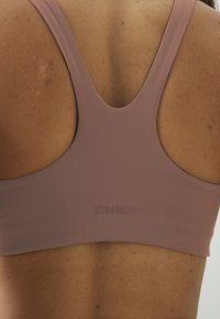 Mauve sportski grudnjak s dizajnom racerback, s glatkom tkaninom i suptilno reljefnim logotipom na leđima. Mekana tekstura i aerodinamičan oblik.