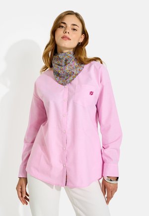Joven mujer vistiendo una camisa rosa claro abotonada sobre una camiseta de cuello alto con estampado floral y pantalones blancos, de pie frente a un fondo blanco.