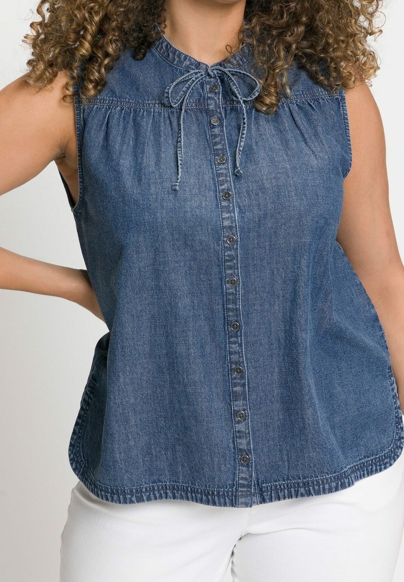Tummansininen denimi ilman hihoja oleva paita, jossa on edessä napit, solmittava kaulus ja kootut olkaset. Ominaisuudet ovat rento malli ja pyöristetty helma.