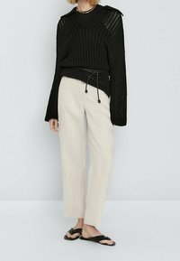 Zwarte gebreide trui met wijde mouwen, hoge halslijn en trekkoorddetail, gecombineerd met lichtbeige broek en zwarte sandalen.