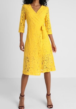 Femme portant une robe portefeuille en dentelle jaune mi-longue avec des manches trois-quarts et une ceinture nouée sur le côté, associée à des talons ouverts foncés.