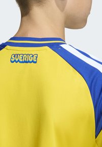 Gul och blå sporttröja med vita ränder, med "SVERIGE"-logotyp i fet, böjd stil på bakre halslinningen. Mjuk textur.