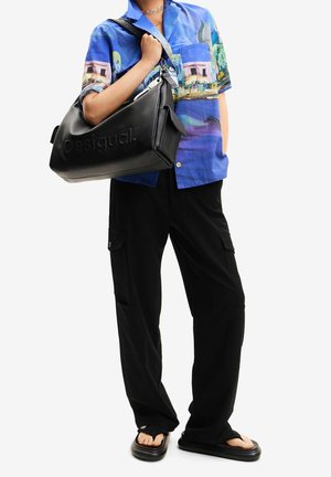 Czarna skórzana torebka z wytłoczonym logo "Desigual", noszona na kolorowej koszulce z krótkim rękawem i luźnych czarnych spodniach cargo. Płaskie sandały.