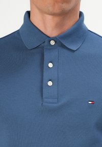 Nahaufnahme einer Person, die ein blaues Polo-Shirt mit drei weißen Knöpfen und einem kleinen rot-weißen-blauen rechteckigen Logo auf der Brust trägt.