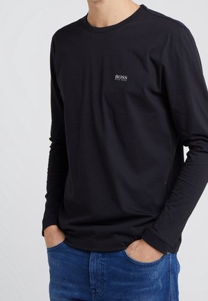 Homme portant un t-shirt noir à manches longues Hugo Boss avec logo sur la poitrine, mains dans les poches, associé à un jean bleu sur fond uni.