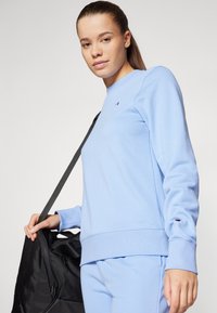 Sudadera azul clara con cuello redondo y puños acanalados, que presenta un pequeño logo en el pecho. El modelo sostiene un bolso de deporte negro.