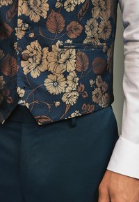Weste mit floralem Muster in Marineblau, verziert mit goldenen und bronzefarbenen Blumen, aus strukturiertem Stoff, mit einer einzelnen Tasche, getragen über dunkelblauen, strukturierten Hosen.