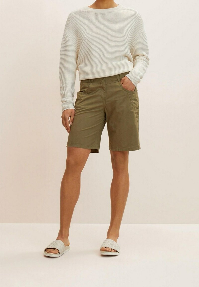 Lichtbeige, gebreide trui met een textuur, losse pasvorm. Olijfgroene shorts met twee zijzakken. Grijze, gewatteerde slidesandalen. Neutrale achtergrond.