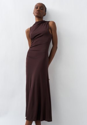 Kobieta w ciemnobrązowej, bezrękawnikowej, długiej sukience z wysokim kołnierzem, stojąca na jednolitym jasnoszarym tle.