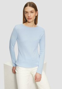 Haut ajusté à manches longues bleu clair avec un col rond, en tissu lisse. Porté avec un pantalon blanc, sur fond neutre.