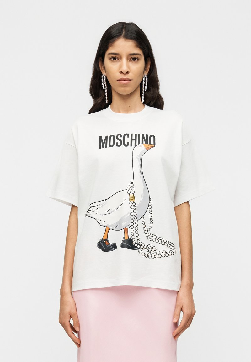 Λευκό βαμβακερό t-shirt με ένα παιχνιδιάρικο γραφικ ό πάπιας με μαύρα παπούτσια που κρατάει ένα νήμα από μαργαριτάρια. Το όνομα της μάρκας "MOSCHINO" είναι τυπωμένο από πάνω.