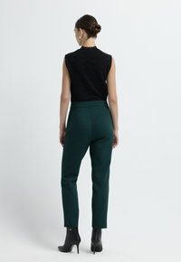 Top nero senza maniche con motivo testurizzato abbinato a pantaloni verde scuro a vita alta e stivali neri alla caviglia. Design semplice e vestibilità snella.