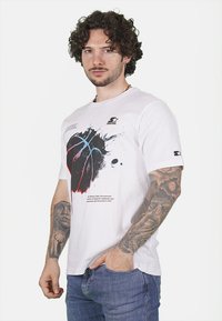 T-shirt bianca in cotone con una grafica di basket con accenti neri, rossi e blu. Collo rotondo e maniche corte con un logo decorativo.