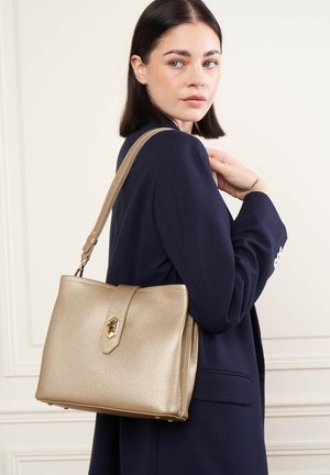 Femme aux cheveux foncés mi-longs portant un blazer bleu marine tient un sac à main en cuir beige texturé avec un fermoir doré sur l'épaule.