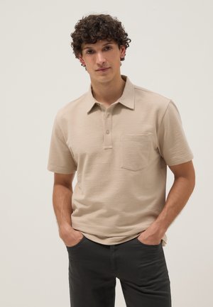 Man met krullend haar die een beige getextureerde korte mouwen polo met borstzak en zwarte broek draagt, handen in zakken, tegen een effen achtergrond.