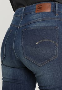 Jeans de mezclilla azul oscuro con una alta cintura, que cuentan con dos bolsillos traseros y una etiqueta de cuero. Textura suave, detalles de costura sutiles.