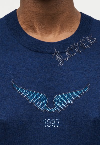 Κοντινή λήψη ενός μπλε πουλόβερ με σχέδιο φτερών από στρας, «1987» κάτω και το όνομα «Players» γραμμένο κοντά στη λαιμόκοψη.