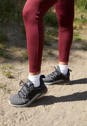 Personne portant des leggings bordeaux et des baskets Adidas grises, debout sur un chemin de terre avec de l’herbe à proximité au soleil.