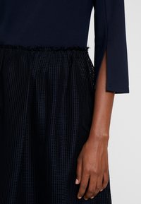 Navy dress with a fitted top and textured, pleated skirt. Features a ruffled hem and side slits on the sleeves. Soft fabric, structured shape.