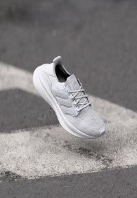 Hellgraue Sportschuh mit einem Mesh-Obermaterial, drei charakteristischen Streifen an der Seite und einer strukturierten Sohle, dargestellt aus einer leichten Aufwärtsneigung.