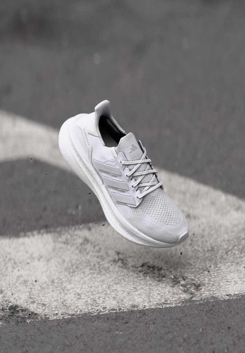 Hellgraue Sportschuh mit einem Mesh-Obermaterial, drei charakteristischen Streifen an der Seite und einer strukturierten Sohle, dargestellt aus einer leichten Aufwärtsneigung.