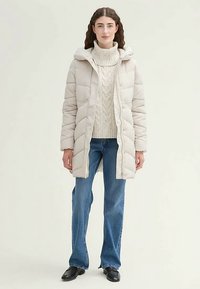 Gepolsterter, cremefarbener Mantel mit Reißverschluss vorne und Kapuze, getragen über einem cremefarbenen, kabelgestrickten Pullover und blauen Schlagjeans. Schwarze Schuhe.