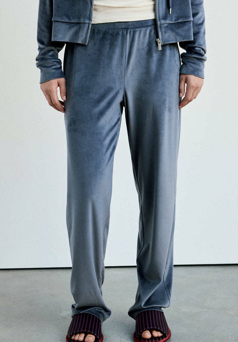 Bas du corps d'une personne portant un pantalon en velours gris et des chaussons ouverts à rayures foncées, debout sur un sol en béton contre un mur clair.